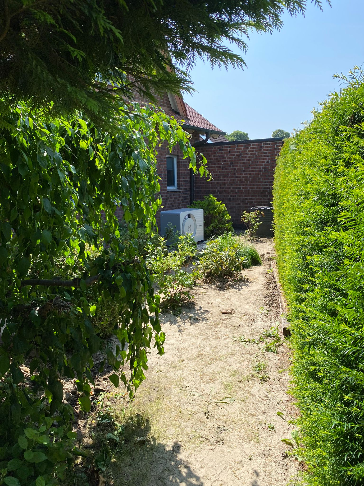 Außengerät einer Wärmepumpe auf Kiesbett vor rotem Ziegelhaus, von Büschen und Bäumen umgeben.